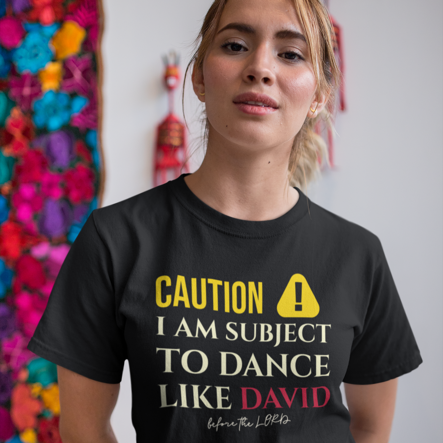 Person wearing a black t-shirt with humorous text design in front of colorful wall art.