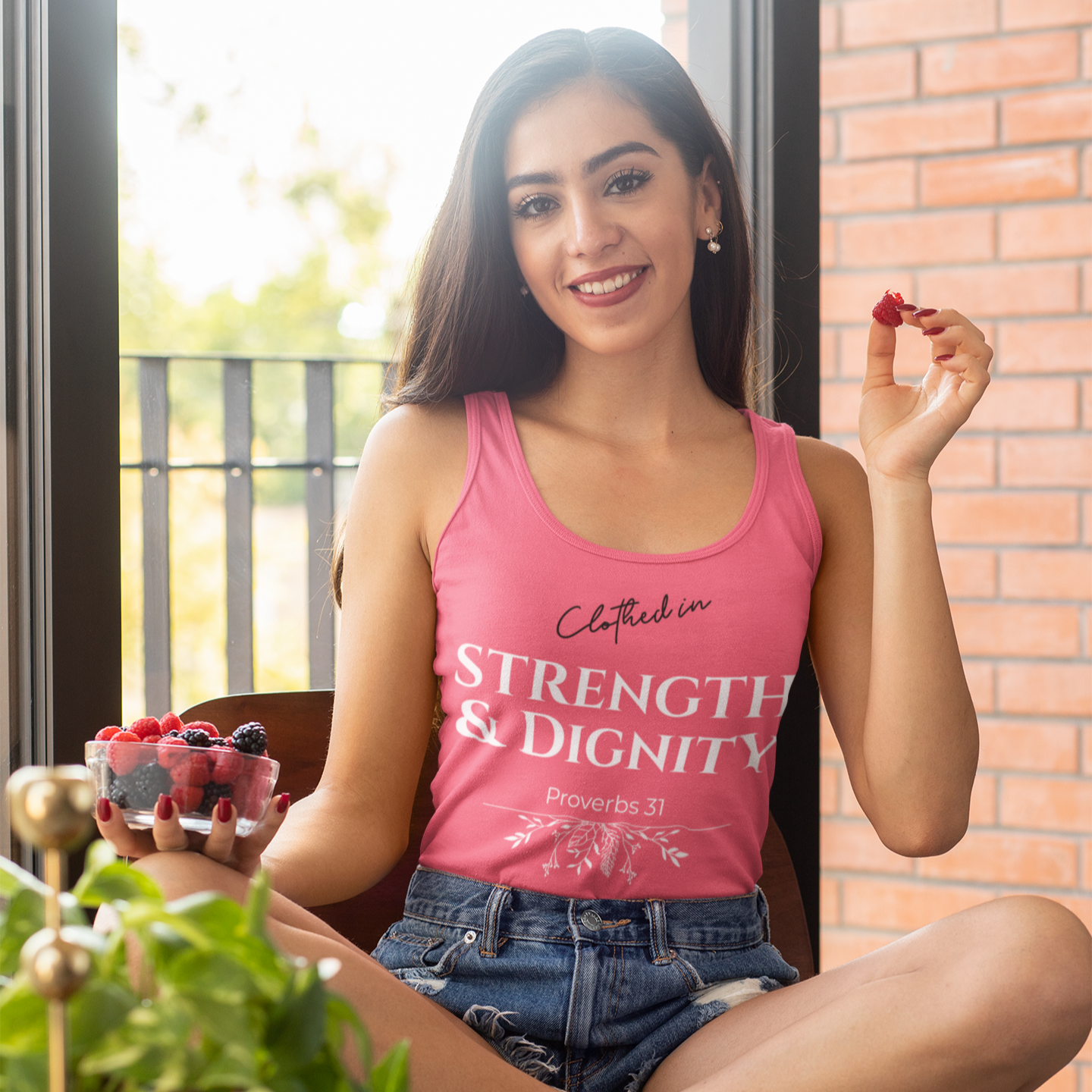 Woman wearing a pink tank top with text, sitting indoors near a window.