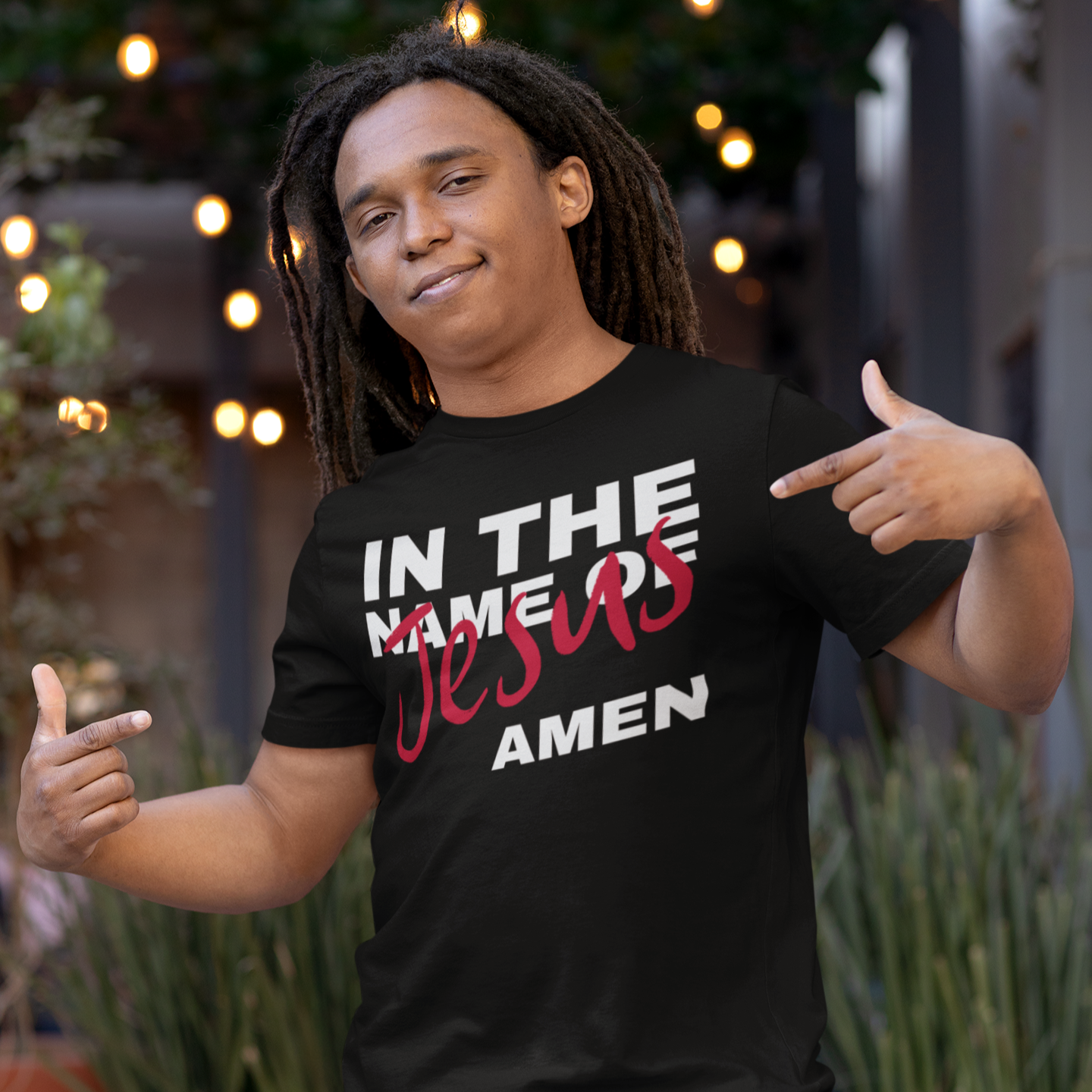 Person wearing a black t-shirt with text, standing outdoors with plants and lights in the background