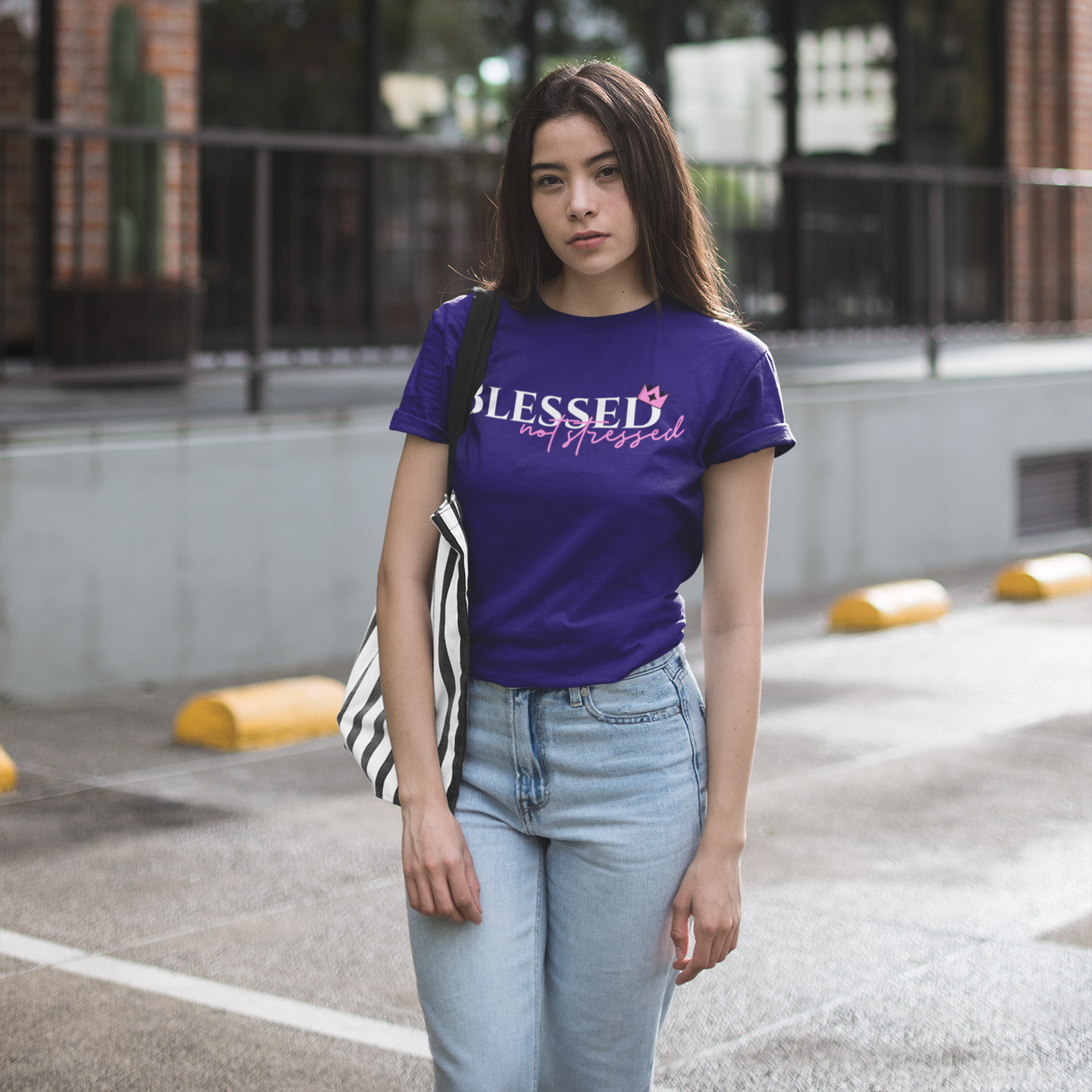 Woman wearing a purple t-shirt with text that says 'blessed not stressed, standing in an urban setting.