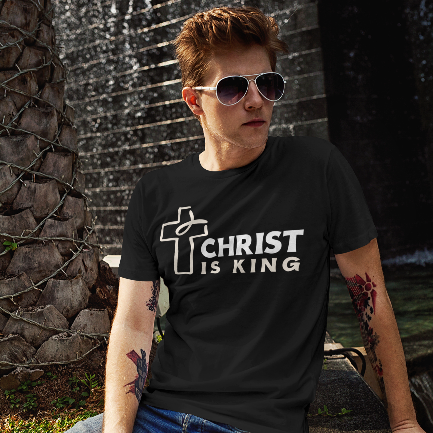 Person wearing a black t-shirt with 'Christ is King' design sitting in front of a water fountain.