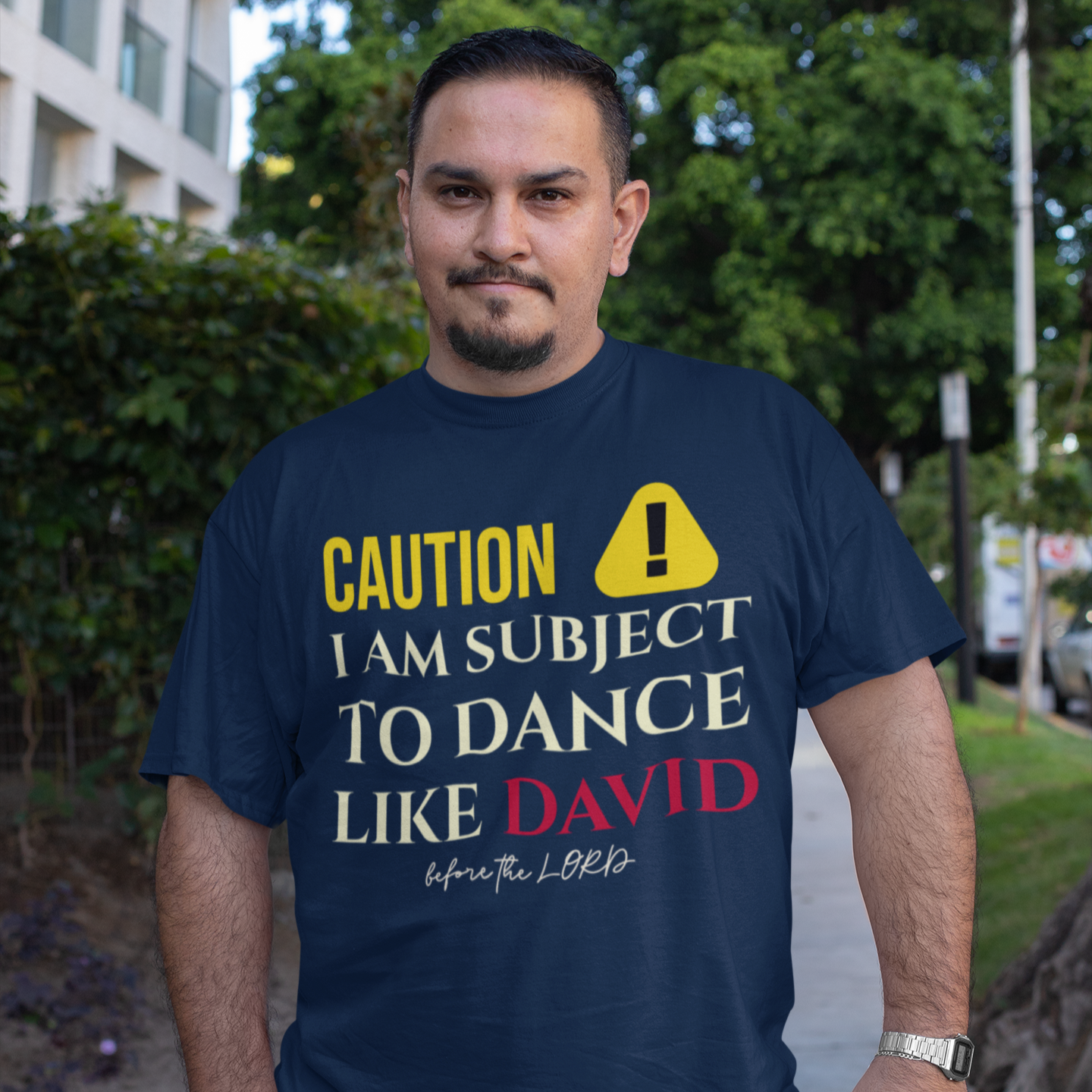 man wearing a navy t-shirt with yellow and red text that says "caution I am subject to dance like david before the lord."