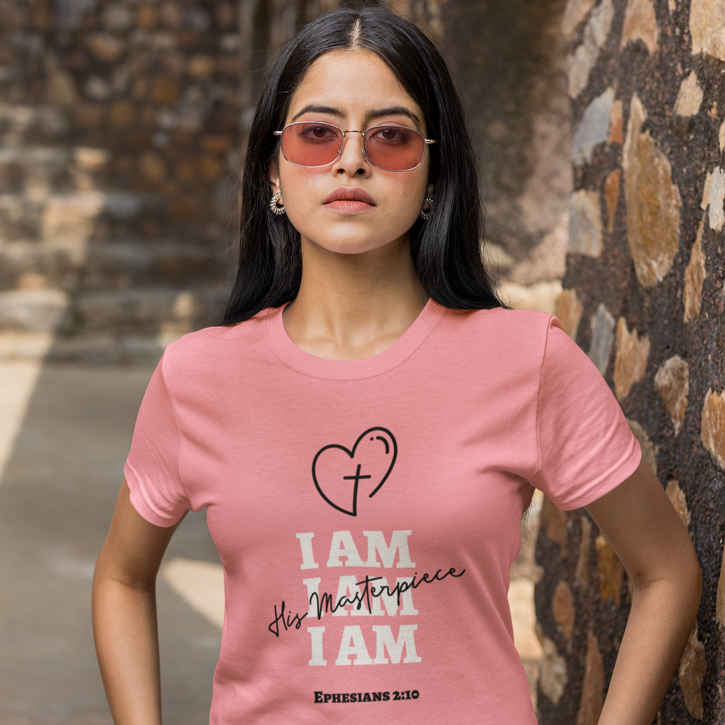 Woman wearing a pink t-shirt with a heart and text design, standing against a stone wall.