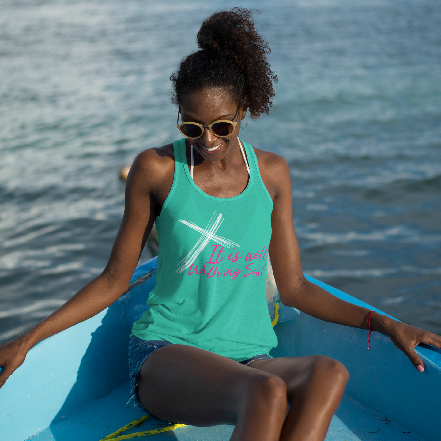 Woman sitting on a blue boat wearing a green tank top with text and sunglasses, with water in the background.