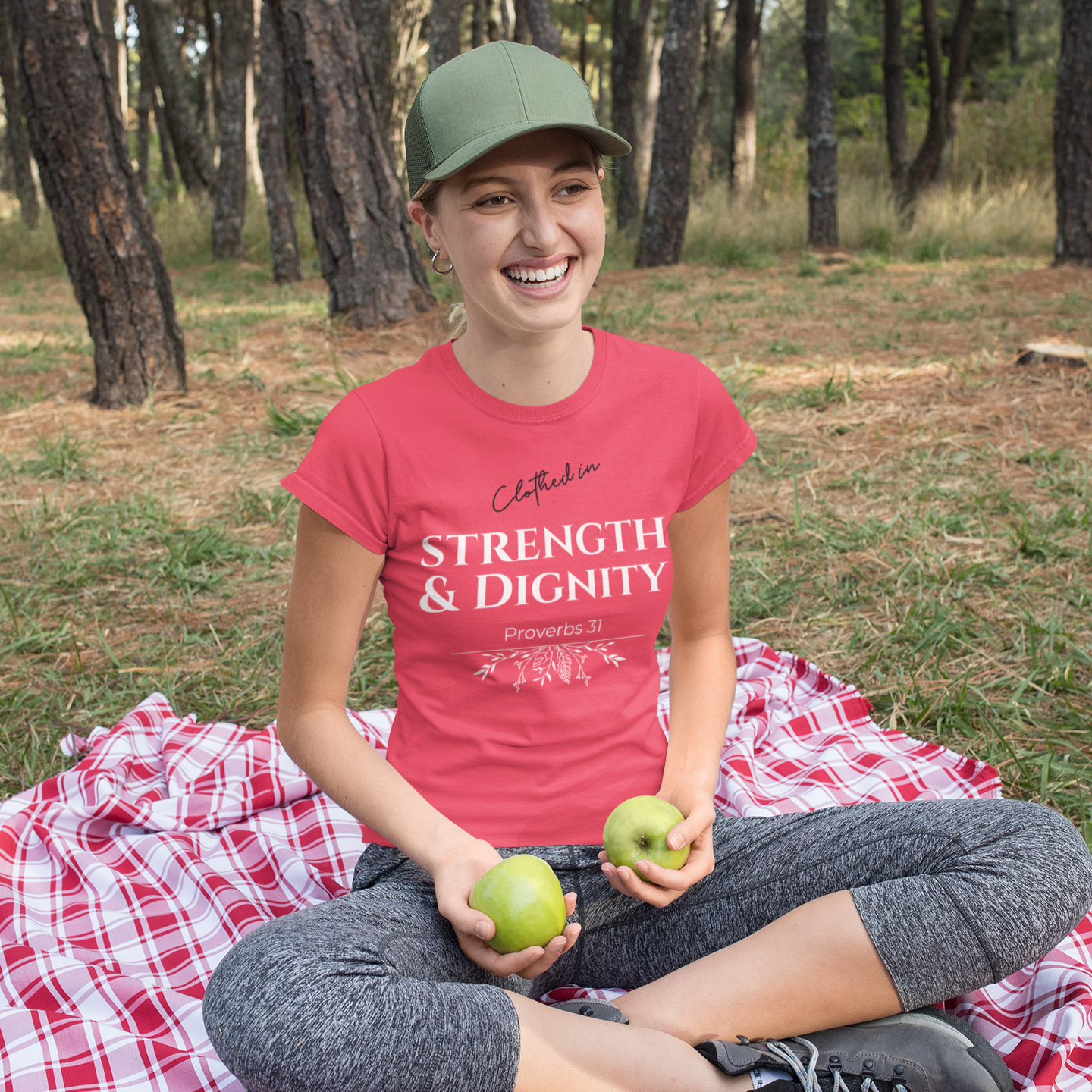 woman sitting on a red and white checkered blanket in a forest, holding apples and wearing a pink t-shirt with 'Clothed in Strength & Dignity Proverbs 31' text